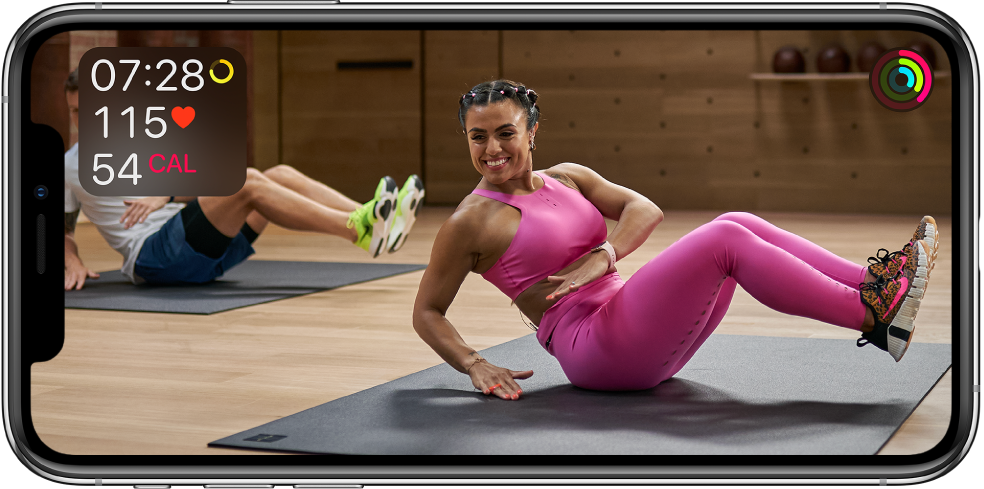 Ekranas, kuriame rodoma trenerio vedama „Apple Fitness “ treniruotė. Informacija apie treniruotės trukmę, pulso dažnį ir sudegintas kalorijas pateikiama viršutiniame kairiajame kampe. Judėjimo, treniruočių ir ant kojų praleisto laiko žiedai pateikiami viršutiniame dešiniajame krašte.