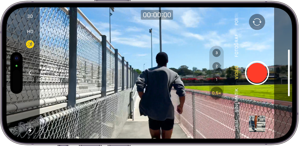 iPhone yatay olarak çevriliyor. Kamera açık ve Video modunda. Flaş ve Hareket modu düğmeleri ekranın sol alt köşesinde görünüyor ve Hareket modu açık. Kamera Denetimleri düğmesi ekranın sol orta kısmında, sol üst köşede ise video çözünürlüğünü ve kare hızını değiştirmek için hızlı anahtarlar var. Sağ tarafta, en üstten en alta doğru Kamera Seçici Arka Yüz düğmesi, Kayıt düğmesi ve Fotoğraf ve Video Görüntüleyici düğmesi yer alıyor. Kamera çerçevesinin içinde koşan bir kişi var.