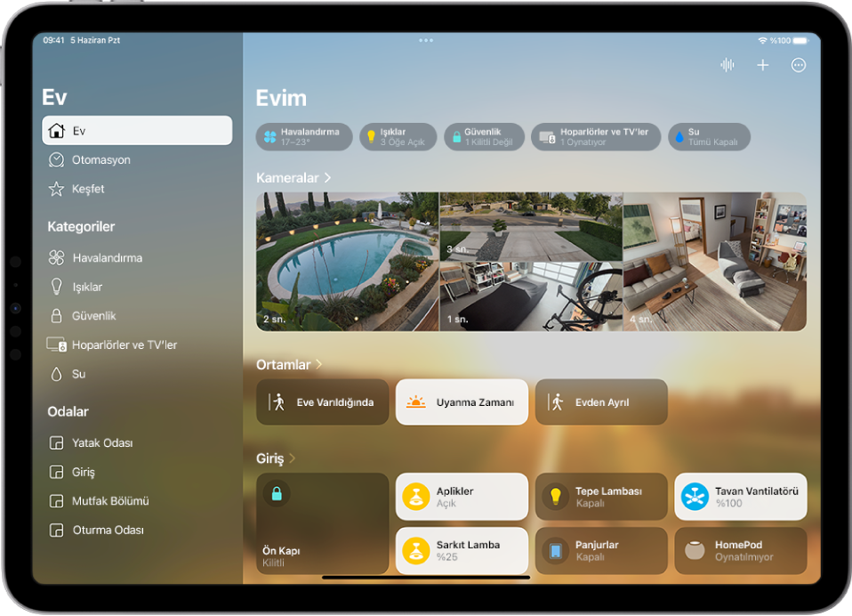 Ev uygulaması, sol tarafta kenar çubuğu ile sol üst tarafta Ev, Otomasyon ve Keşfet sekmelerini gösteriyor. Evdeki Kategoriler ve Odalar bölümleri alt tarafta görünüyor. Kenar çubuğunun sağında, en üstte İnterkom, Ekle ve Daha Fazla Seçenek düğmeleri bulunuyor. Kategori düğmeleri üst tarafta görünüyor. Onun altında ortamlar, kamera görüntüleri ve içinde aksesuarlarıyla bir oda var.