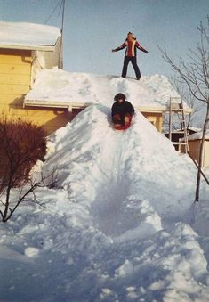 This may contain: a man riding a snowboard down the side of a pile of snow next to a house