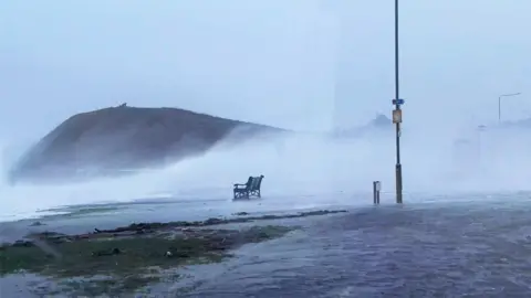 Waves crash onto a road in Troon, with water sweeping over a bench