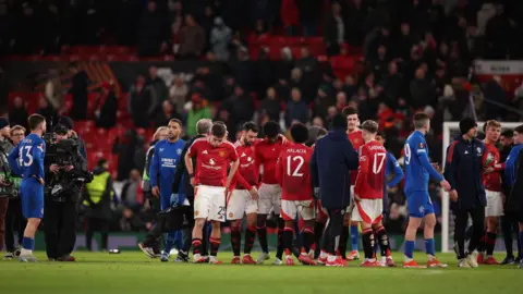 Manchester United and Rangers players on the Old Trafford pitch after the match - the Rangers players, in all blue kits, look dejected with hands on hips, while the Manchester United players - in red shirts and white shorts look tired, some with their arms around each other