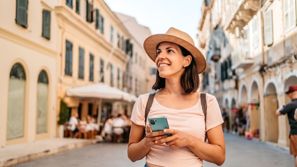 Junge Touristin wandert mit Sonnenhut und Smartphone durch eine belebte mediterrane Straße