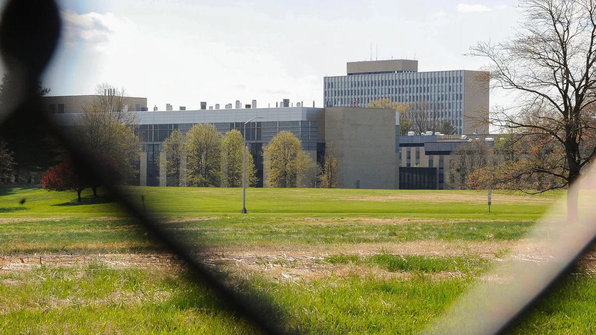 A large commercial building with a green grass lawn is framed through a wire metal fence mesh. National Institutes of Standards and Technology in Gaithersburg, Md., on April 17, 2022.