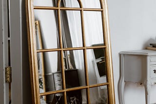 A gold mirror with an arched top and many facets within it leans against a white wall in an apartment setting.