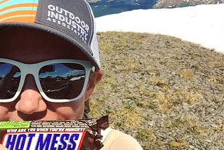 Author standing on mountain top, holding a Snickers bar that says “hot mess” on label