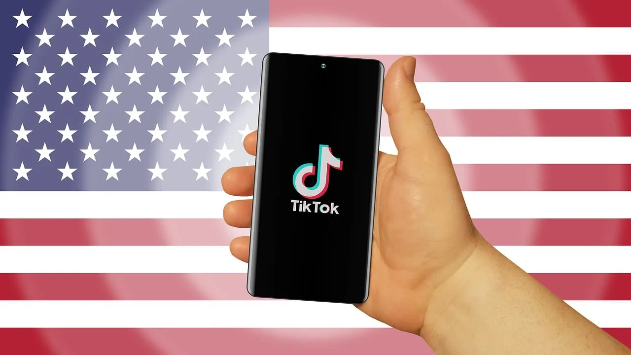 IMAGE: A hand holding a smartphone with the TikTok logo on it, on a background with the US flag