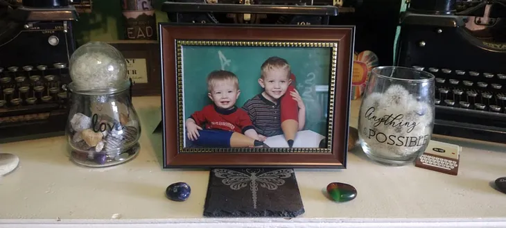 Photo of a 3-year-old and five-year-old boy each holding giant blue and red pencils sitting in front of a chalkboard. This photo is sitting between two glasses one labeled with Love and one labeled with Anything is Possible. There is a dragonfly coaster and two heart rocks in the foreground.