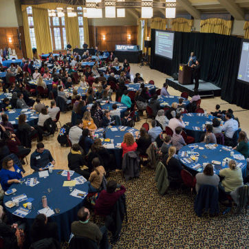 Large gathering of people sitting at round tables listening to a presentation