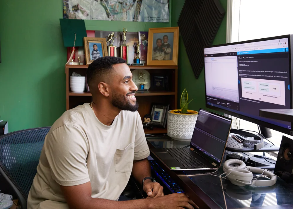 Karrim Omer, a Google Data Analytics certificate graduate, sits on a chair in a bedroom and is smiling at a computer.