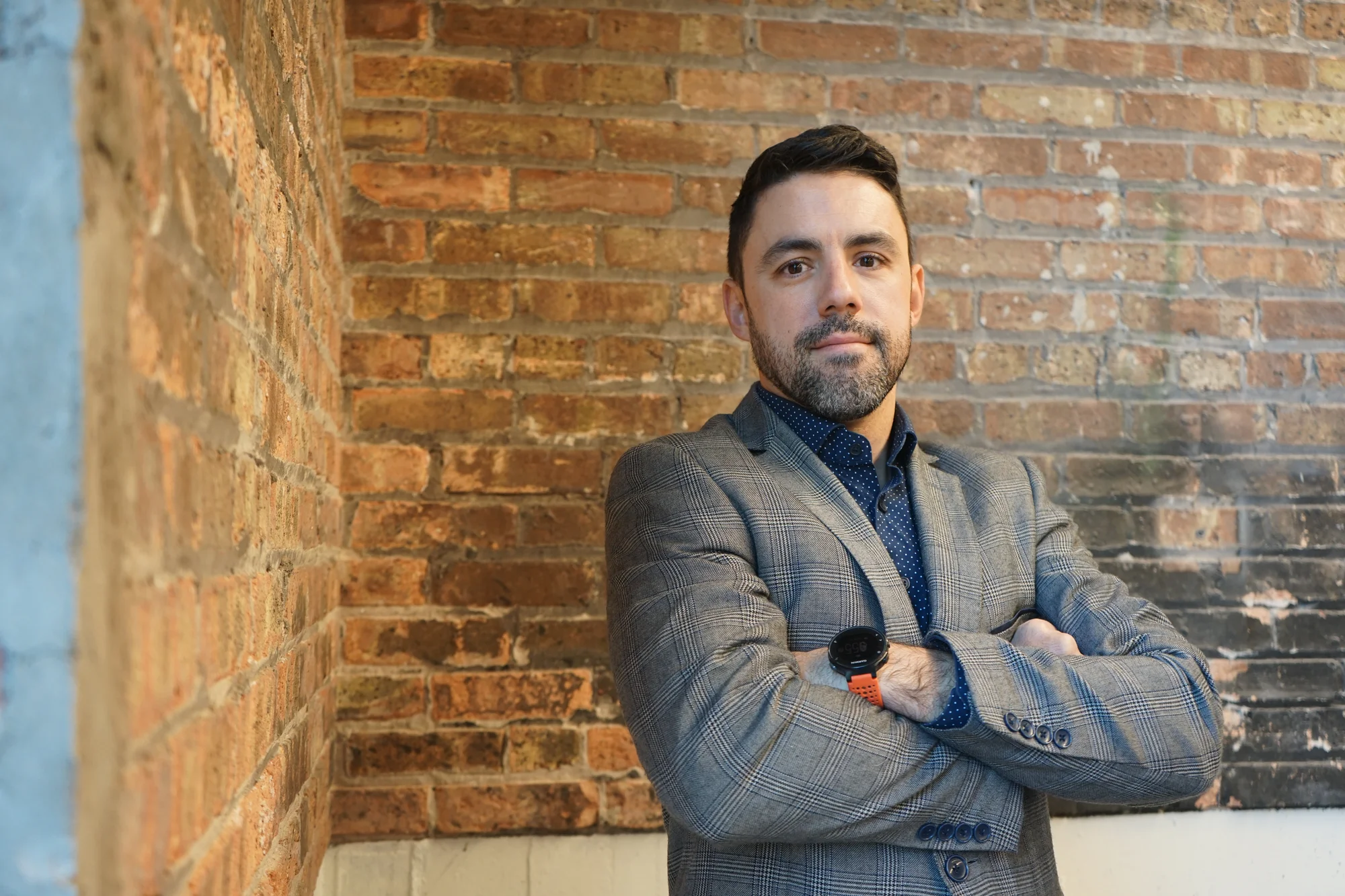 A man wearing a gray suit stands with his arms crossed in front of a brick wall.