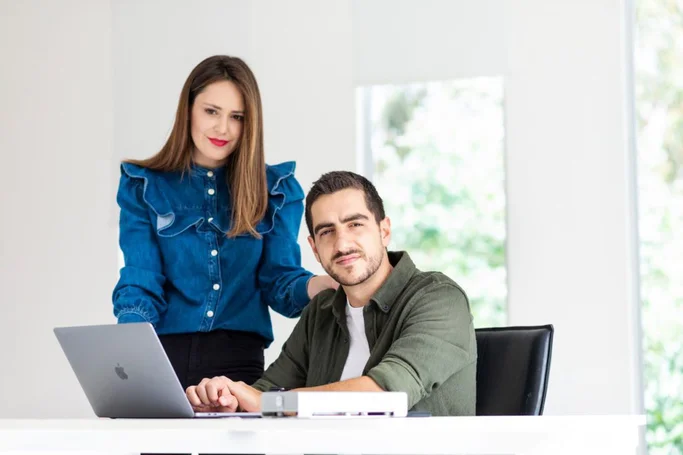 A woman wearing a blue shirt stands next to a man sitting in a chair behind a laptop.