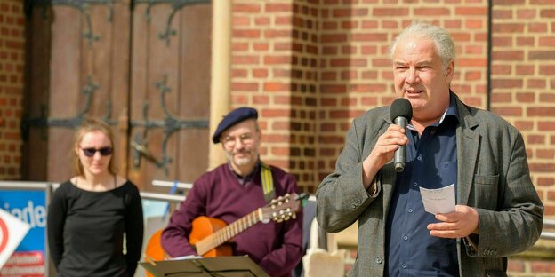 Andrej Hunko auf der Kundgebung beim Ostermarsch in Herne, er hält ein Mikrofon in der Hand. Hinter ihm steht ein Mann mit Gitarre vor einem Notenständer, daneben eine junge Frau mit Sonnenbrille