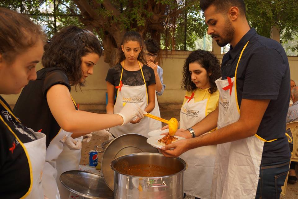 People stand over a large pot of stew, as a woman serves a plate of food with help from a colleague.