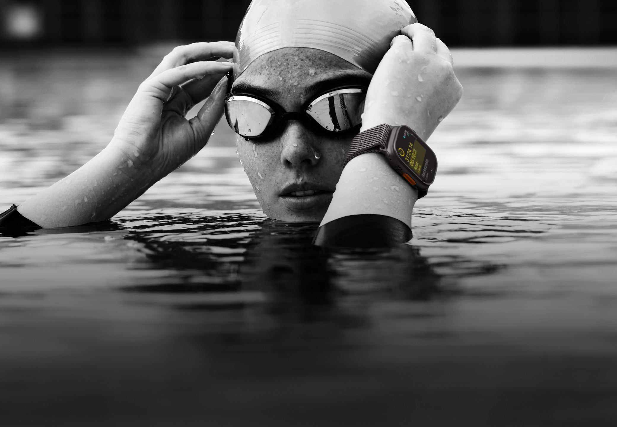 A swimmer's head and hands above water in a swimming pool wearing Apple Watch Ultra 2 and adjusting goggles.