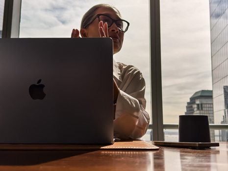 Egy nő asztalnál ül egy sokemeletes épületben. A mögötte lévő nagy ablakon keresztül más épületek látszanak. A nő előtt egy nyitott MacBook van.