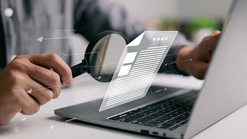 A person at a laptop examining the screen with a magnifying glass.