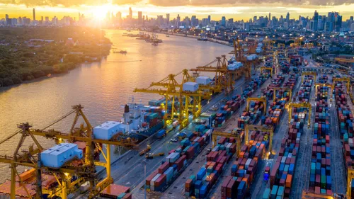 Photo of a cargo shipping port at sunset showing cranes, water docking access, and large amounts of cargo containers.