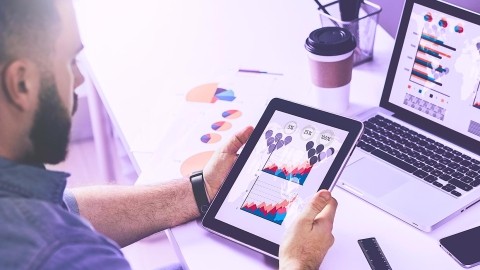 Image of man holding tablet with graphs, in front of laptop with more graphs