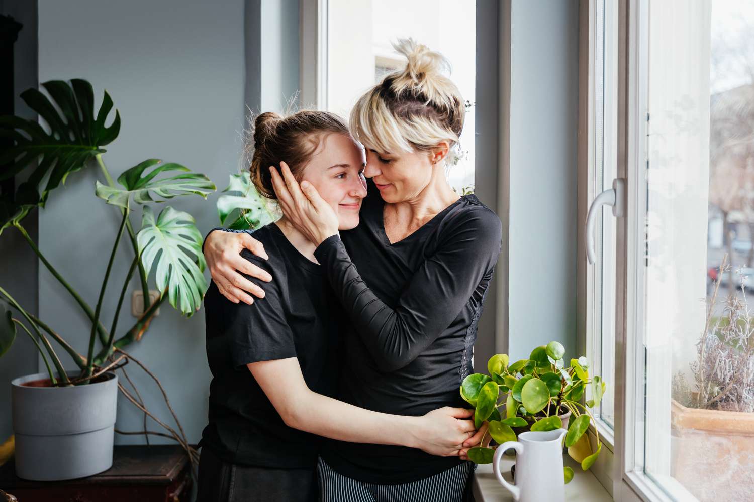 A mom and a daughter hug near a window