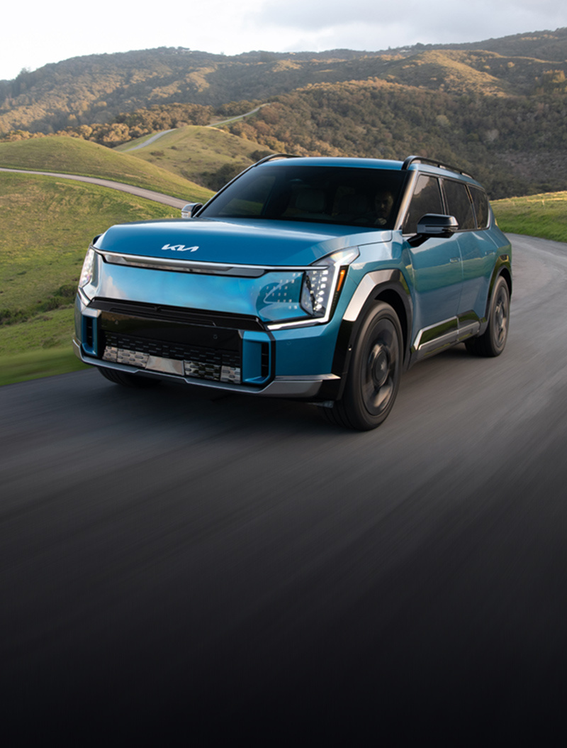 2024 Kia EV9 in blue, three-quarter front view, driving through a paved road in the hills