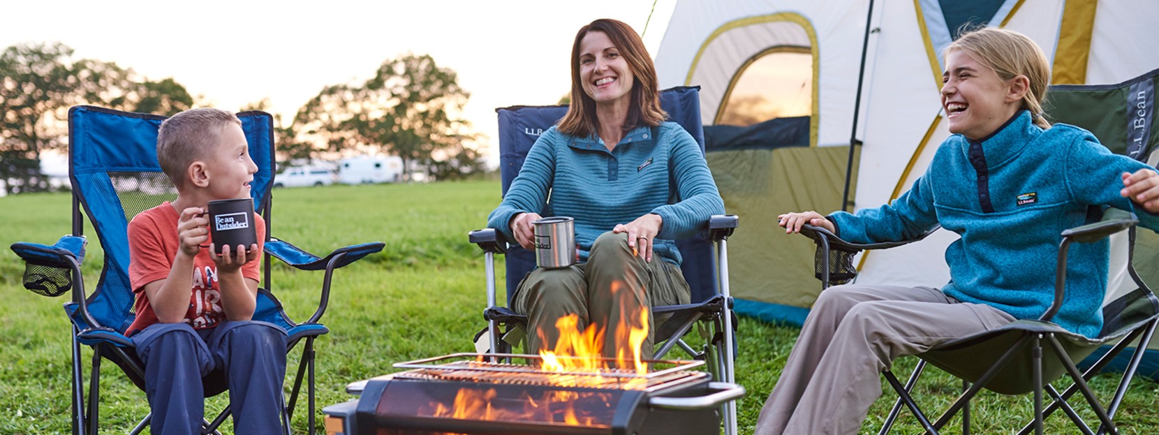 A woman and two children sit by a fire in front of a tent