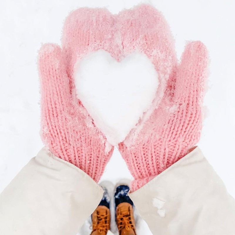 A person with pink mittens holding snow in the shape of a heart