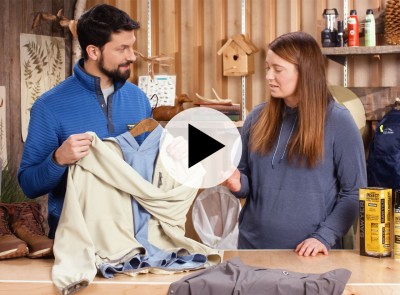 A man and a woman looking at an insect repelling shirt, a play video icon in the center.