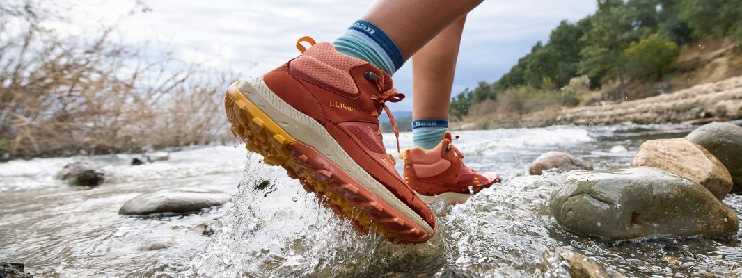 A close-up of feet in hiking boots walking through a rocky shallow stream.