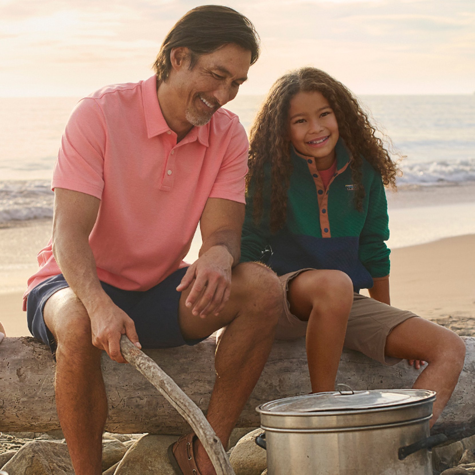 Father and daughter sitting on a log on the beach by a campfire.