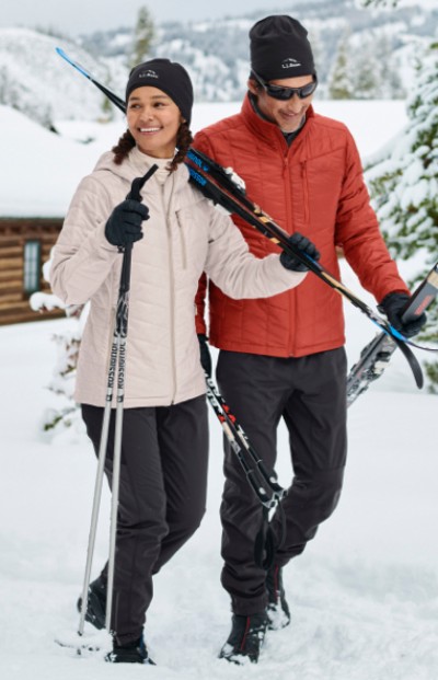 Two individuals with ski equipment stand in the snow, ready for winter sports, with a snowy landscape in the background.