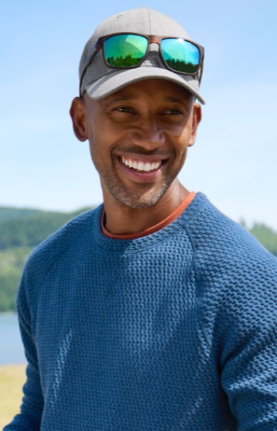 A man in a blue sweater and cap stands before a blurred natural backdrop.