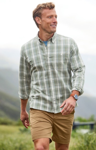 A man wearing a checkered shirt, shorts, and a watch, stands outdoors with mountains in the background.