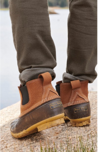 A man wearing L L Bean boots stands on a sandy shore, with a serene lake and distant land in the backdrop.