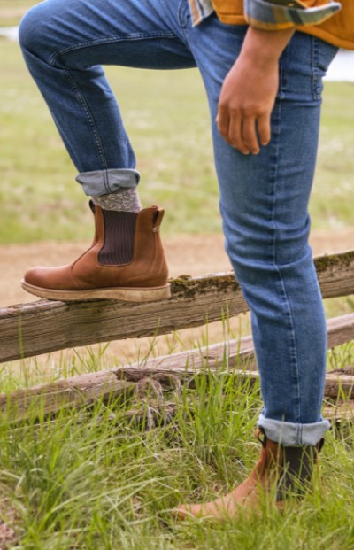 A man in jeans and brown boots steps over a wooden fence in a grassy field, capturing a moment of rustic adventure and freedom.