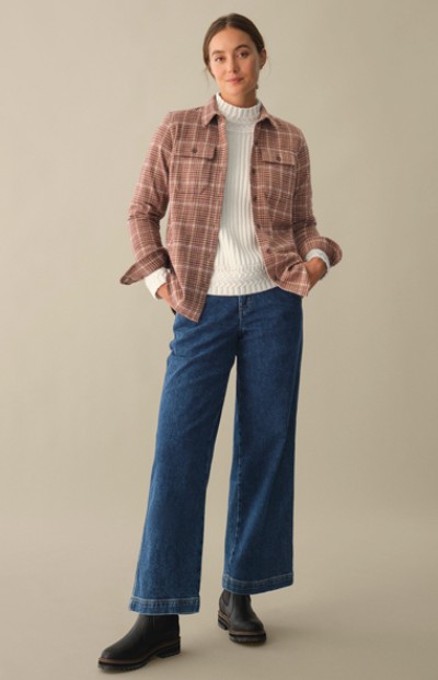 A woman in a plaid jacket, white sweater, and blue jeans is casually standing indoors.