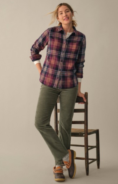 A woman stands with one foot on a chair, wearing a plaid shirt and green pants. The scene is casual and indoors. 