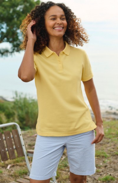 A woman wearing a yellow polo and blue shorts stands along side a lake.