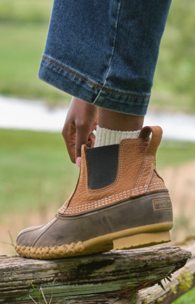 A close up of a person’s foot in a brown L L Bean Boot, standing on a wooden beam, with greenery in the background.