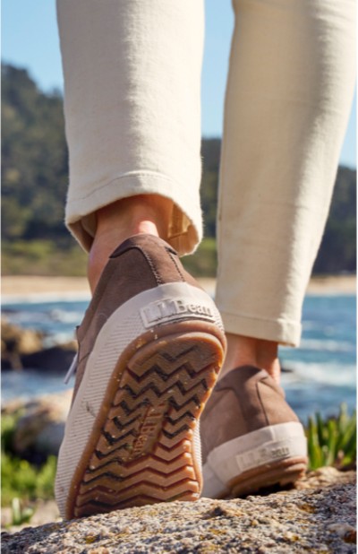 A person in beige pants and brown sneakers walks on a sandy path with water and rocks in the background.