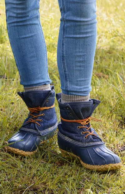 A woman stands on grass, wearing blue L L Bean boots with orange laces on grass.