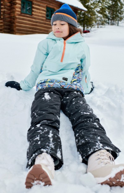 A child sits in the snow, wearing snow pants outside a log cabin. The scene is serene and wintry. 