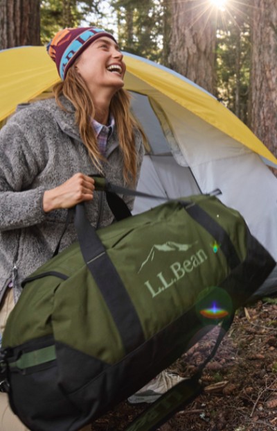 A woman is setting up a campsite, holding a large L L Bean duffel bag near an open tent in a forest.