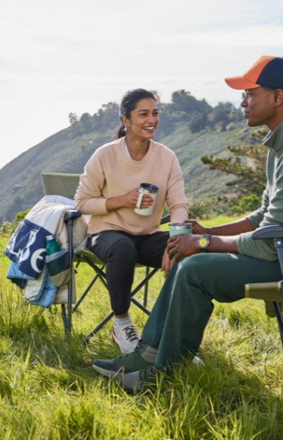 Two people are seated outdoors on L L Bean Camp Comfort Chairs, with a scenic hilly backdrop, enjoying a moment in nature.