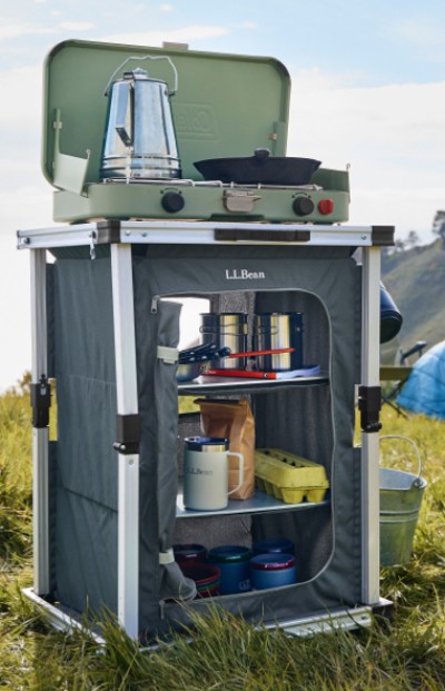 Portable outdoor kitchen setup with stove, supplies, and L L Bean branding in a grassy field. 