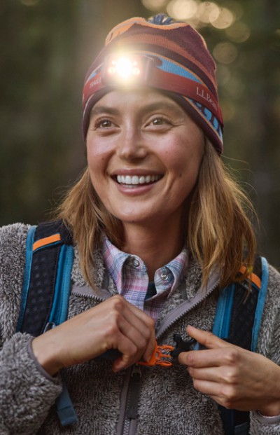 A woman wearing a headlamp and L L Bean backpack, face obscured, focusing on the zipper of a jacket, suggesting preparation for an outdoor activity.