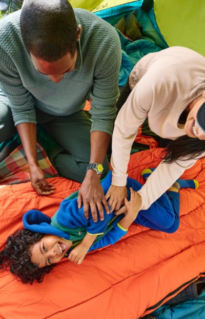 Two adults attend to a child lying on an orange sleeping bag inside a tent, creating a fun scene during a camping trip.