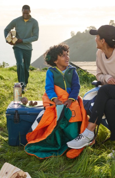 Three people enjoy a picnic. One holds a book, another sits in an orange sleeping bag, and the third sits beside a cooler.