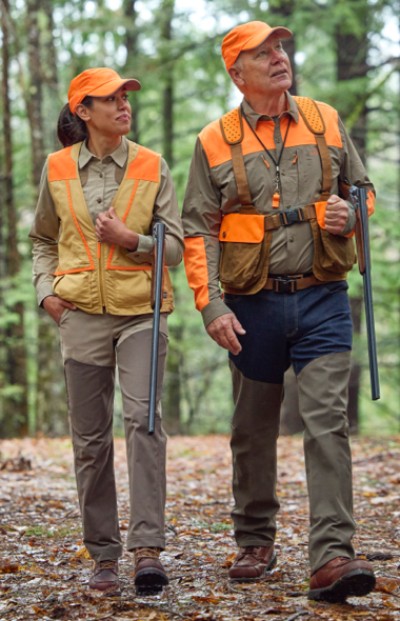 Two hunters in orange vests and hats quietly walk through a dense forest in search of wildlife. 