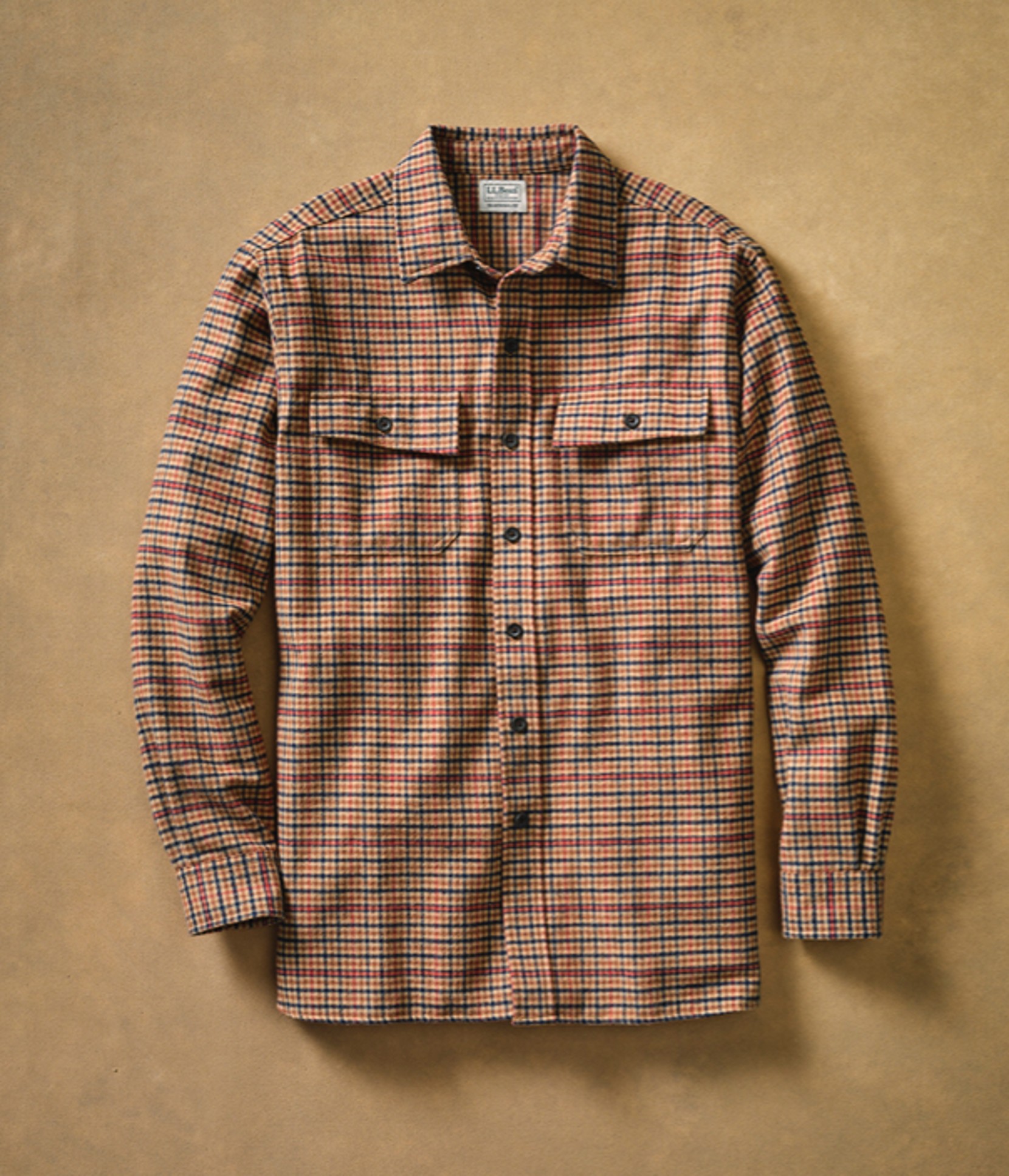 A plaid L L Bean shirt with a collar, buttons, and pockets is neatly laid out on a beige background.